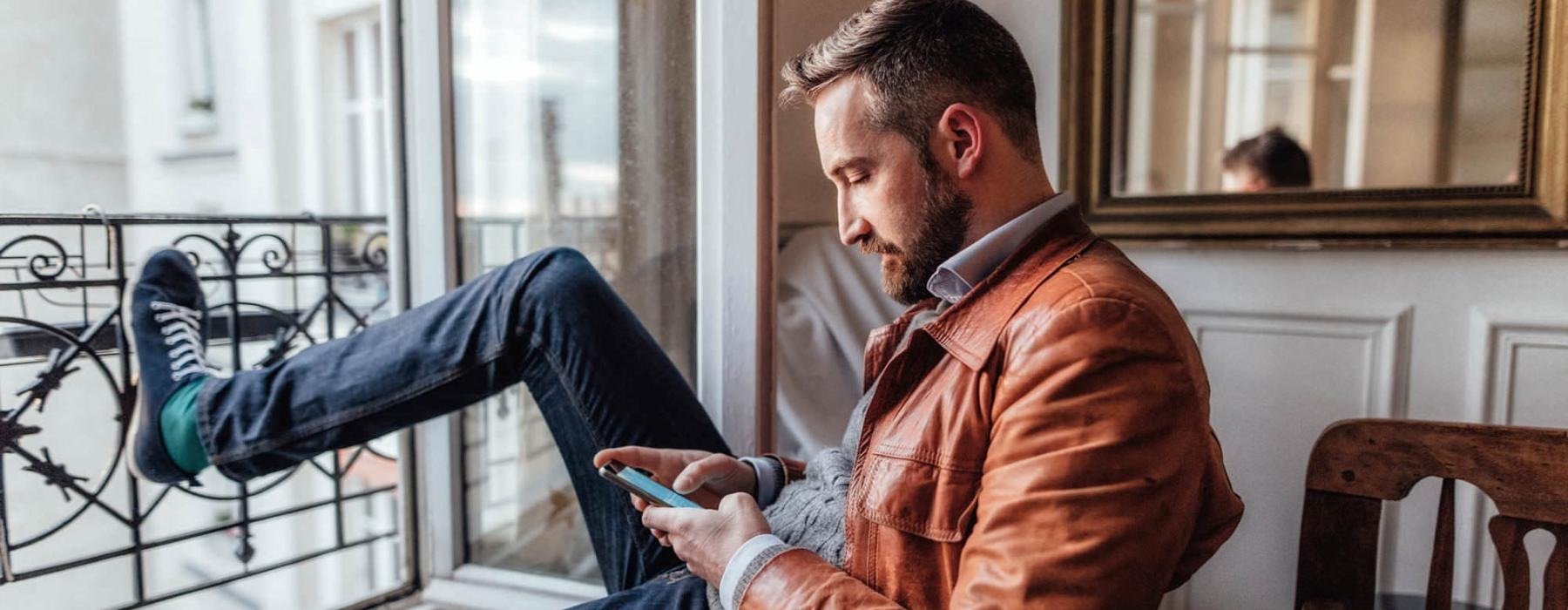 a man sits on a chair and rests his foot on a balcony railing as he looks at his phone in his room